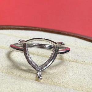 May include: A silver ring with a shield-shaped setting. The ring features a slender band and a pronged setting designed to hold a gemstone. The ring is displayed on a light-coloured surface.