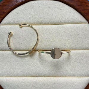 Puede incluir: Dos anillos dorados. Uno es una banda fina con un pequeño círculo plano. El otro es un anillo abierto curvado con pequeños extremos de bola. Ambos anillos se muestran sobre una superficie de color crema.