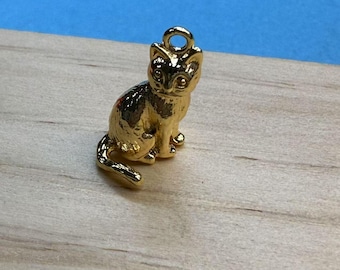 May include: A gold-colored cat charm with a loop at the top for attaching to a necklace or bracelet. The cat is sitting with its tail curled and has detailed facial features. The charm is on a light wood surface.
