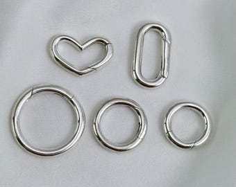May include: A collection of five silver-toned metal spring gate rings and clasps in various shapes, including one heart-shaped clasp, one elongated oval clasp, and three circular rings of different sizes, each featuring a functional spring mechanism.