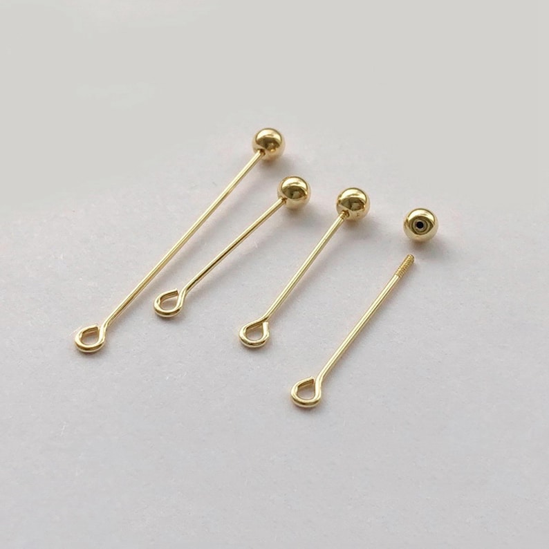 Peut inclure: Plusieurs appr&ecirc;ts de bijoux dor&eacute;s, dont des &eacute;pingles droites avec une petite boucle &agrave; une extr&eacute;mit&eacute; et une perle sph&eacute;rique &agrave; l'autre. Une &eacute;pingle a une extr&eacute;mit&eacute; filet&eacute;e. Les appr&ecirc;ts sont dispos&eacute;s sur une surface de couleur claire.