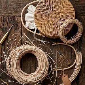Könnte beinhalten: Eine Sammlung von Korbweide-Materialien, darunter ein runder Korb, Spulen aus naturfarbenem Rattan, ein hölzerner Stickrahmen und kleine Werkzeuge. Die Gegenstände sind auf einer dunklen Holzoberfläche angeordnet, was auf ein Bastel- oder DIY-Projekt hindeutet.