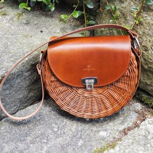 May include: A round, woven wicker handbag with a brown leather flap and strap. The bag has a silver clasp and metal rings. The leather flap is embossed with the word "ANGELIE". The bag is sitting on a stone surface.