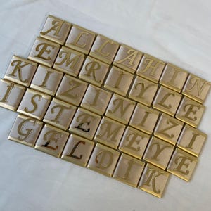 Könnte beinhalten: Goldfarbene Buchstabenkacheln, die den Satz "ALLAHIN IZNIYLE GEL DIK" auf einem hellbeigen Hintergrund buchstabieren.