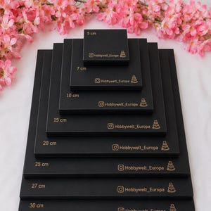 Puede incluir: Una pila de bases cuadradas negras para pasteles de varios tamaños, de 5 cm a 30 cm. Cada base tiene el nombre de Instagram "Hobbywelt_Europa" y un icono de pastel dorado. Flores rosas en el fondo.
