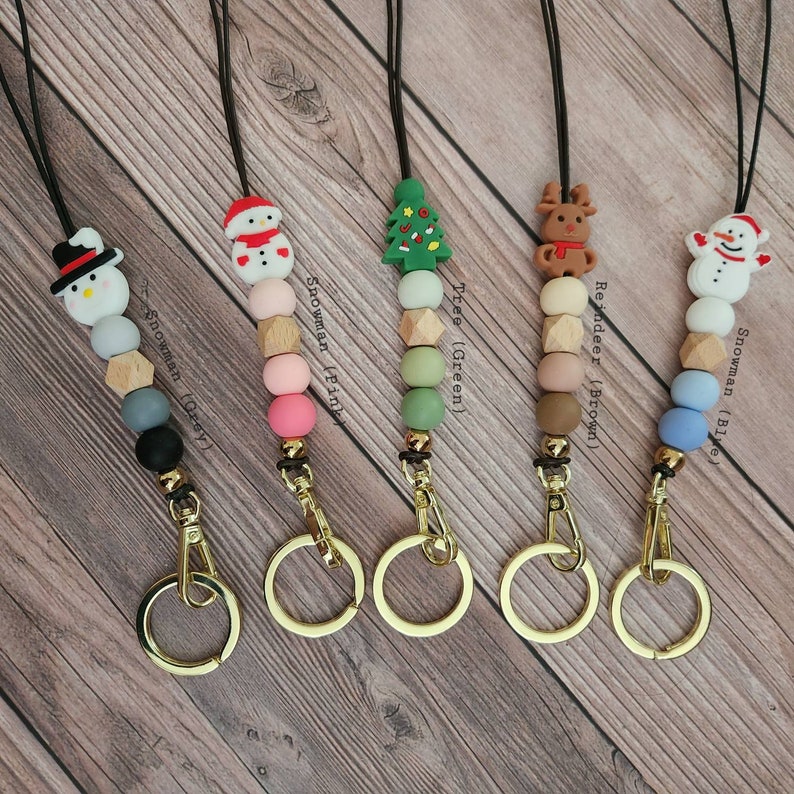 May include: A collection of five Christmas-themed lanyards. Each lanyard features a different design: a snowman (gray), a snowman (pink), a green tree, a brown reindeer, and a snowman (blue). The lanyards have gold-colored clasps and key rings.