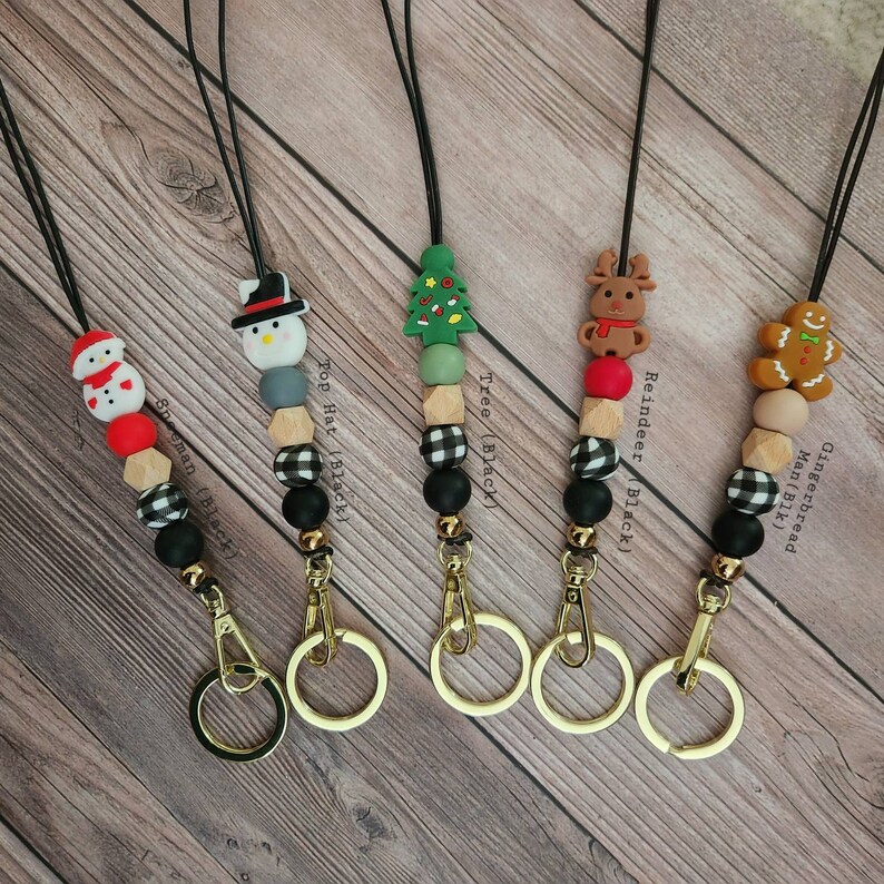 May include: Christmas-themed lanyards with various charms, including a snowman, Christmas tree, reindeer, and gingerbread man. Each lanyard features a black cord, gold-colored hardware, and a mix of black, white, and wooden beads. Text on the image reads "Snowman (Black)", "Top Hat (Black)", "Tree (Black)", "Reindeer (Black)", and "Gingerbread Man (Blk)".