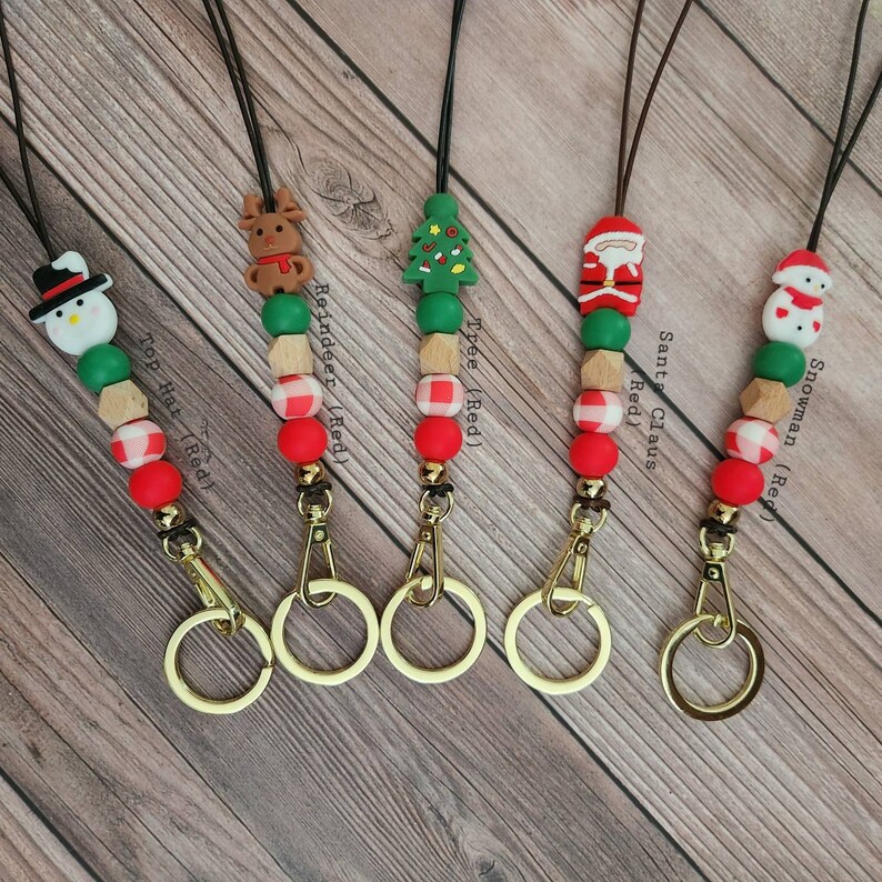 May include: Five Christmas-themed lanyards with black cords, gold-colored clasps, and various bead designs. Each lanyard features a different holiday figure: a snowman, reindeer, Christmas tree, Santa Claus, and a snowman. The beads are red, green, and natural wood-toned.