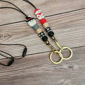 May include: Two beaded lanyards with gold-colored key rings. One lanyard features a snowman with a black hat, the other a snowman with a red hat. The beads are black, gray, red, and wood-toned, with some in a buffalo plaid pattern.