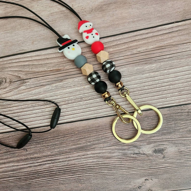 May include: Two beaded lanyards with gold-colored key rings. One lanyard features a snowman with a black hat, the other a snowman with a red hat. The beads are black, gray, red, and wood-toned, with some in a buffalo plaid pattern.