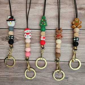 May include: Five Christmas-themed lanyards with gold-colored clasps. Each lanyard features a different holiday-themed charm, including a snowman, Christmas tree, reindeer, and gingerbread man. The lanyards are made with a combination of wooden and silicone beads.