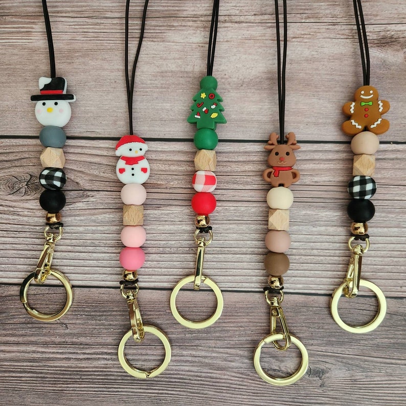 May include: Five Christmas-themed lanyards with gold-colored clasps. Each lanyard features a different holiday-themed charm, including a snowman, Christmas tree, reindeer, and gingerbread man. The lanyards are made with a combination of wooden and silicone beads.