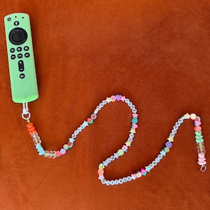 Könnte beinhalten: Eine limettengrüne TV-Fernbedienung mit einem Perlenband. Das Band zeigt weiße Buchstabenperlen mit der Aufschrift "I WILL TAKE YOU TO MY COTTAGE THIS SUMMER", die mit bunten Obst- und sternförmigen Perlen durchsetzt sind. Die Fernbedienung liegt auf einer orangefarbenen Oberfläche.