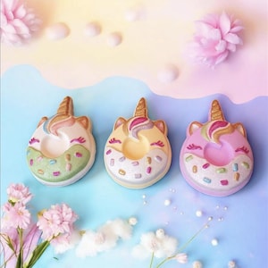 May include: Three unicorn-shaped bath bombs in pastel shades. Each has a gold horn, a colourful mane, and a sprinkle-covered base. The background is a gradient of blue, yellow, and pink with floral accents.