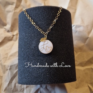 Könnte beinhalten: Eine goldene Kettenkette mit einem weißen runden Anhänger, der ein goldenes Blatt und einen kursiv geschriebenen Buchstaben L mit einem Herzen darin zeigt. Der Anhänger befindet sich auf einem schwarzen Präsentationsständer. "Handmade with Love" steht auf dem Ständer.