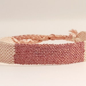 May include: A woven friendship bracelet with a pink and white color scheme. The bracelet is made of thin cords that are woven together to create a pattern.
