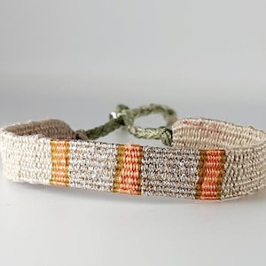 Könnte beinhalten: Ein gewebtes Armband mit einer weißen Basis und Streifen in Orange und Gold.