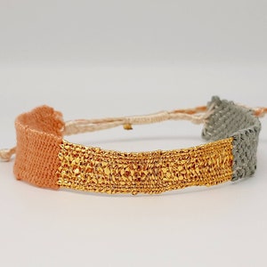 May include: A woven bracelet with three bands of color: peach, gold, and gray. The gold band is made of a textured metal.