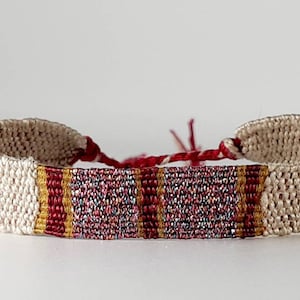 May include: A woven friendship bracelet with a white base and a red, gold, and silver stripe pattern.