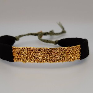 Puede incluir: Un brazalete de oro con un diseño texturizado y extremos de tela negra. El brazalete está atado con un cordón verde.