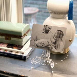 May include: A clear acrylic photo frame with a scalloped edge and a stand. The frame holds a black and white photo of a family with a baby. The frame is engraved with the text "I love you, Mama".