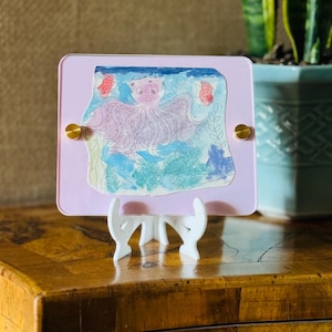 Op de afbeelding: Een paarse octopus met roze accenten is getekend op een wit papier met blauwe, groene en roze aquarelwashes. Het papier is ingelijst in een lichtpaarse acryllijst met gouden accenten. De lijst staat op een witte standaard.