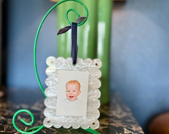 May include: A white scalloped picture frame with a photo of a baby inside. The frame is hanging from a green metal stand with a black ribbon.
