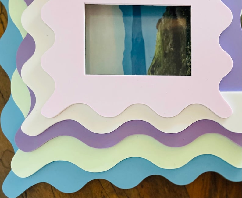 May include: A set of four layered picture frames with wavy edges. The frames are in shades of blue, green, purple, and white. The top frame has a photo of a landscape with a body of water and a rocky shore.
