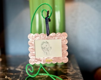May include: A pink scalloped picture frame with a black ribbon hanging from the top. The frame is holding a black and white photo of a young girl. The frame is sitting on a green metal stand. The year 2024 is written in gold on the bottom of the frame.