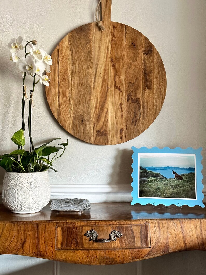 May include: A wooden cutting board with a handle hangs on a white wall above a wooden table with a drawer. A white ceramic planter with a white orchid plant sits on the table. A blue picture frame with a photo of a brown animal in a field sits on the table.