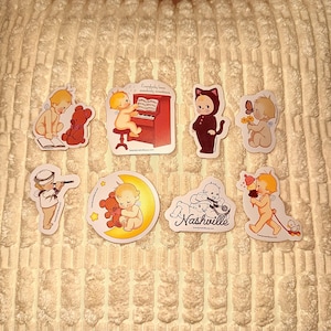 May include: A collection of stickers featuring vintage-style illustrations. The stickers depict cherubic figures, teddy bears, a cat in a costume, and a sailor. Some stickers include text such as "Nashville" and text above a piano.