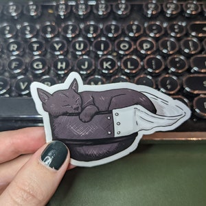 May include: A black and white sticker of a cat sleeping in a helmet. The cat is curled up in the helmet, with its tail wrapped around its body. The helmet has a white strap and a few rivets.