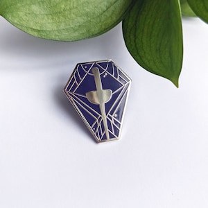 Könnte beinhalten: Eine silberne und blaue Emaille-Anstecknadel mit einem geometrischen Design und einem stilisierten Schwert in der Mitte.