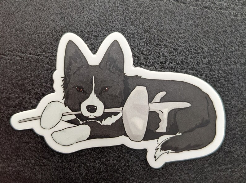 May include: A black and white border collie dog sticker with brown eyes, lying down and holding a fencing foil in its mouth.