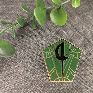 Puede incluir: Un pin de esmalte verde y dorado con una silueta negra de una varita dentro de un diseño geométrico. El pin tiene la forma de un pentágono.