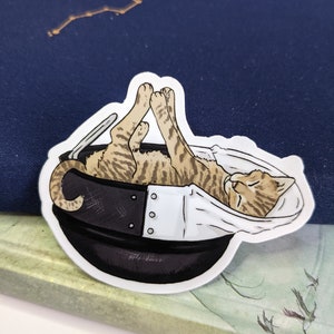 May include: A sticker of a tabby cat curled up inside a black and white pot. The cat is sleeping with its eyes closed. The sticker is on a blue background.