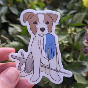 May include: A white and brown dog with a blue glove in its mouth. The dog is sitting on a stick with a grey fish behind it.
