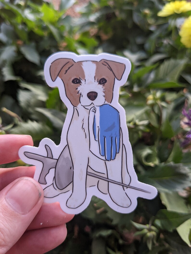 May include: A white and brown dog with a blue glove in its mouth. The dog is sitting on a stick with a grey fish behind it.