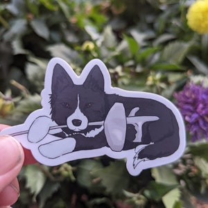 May include: A black and white border collie dog sticker with a fencing foil in its mouth. The dog is lying down and the foil is pointing to the right.