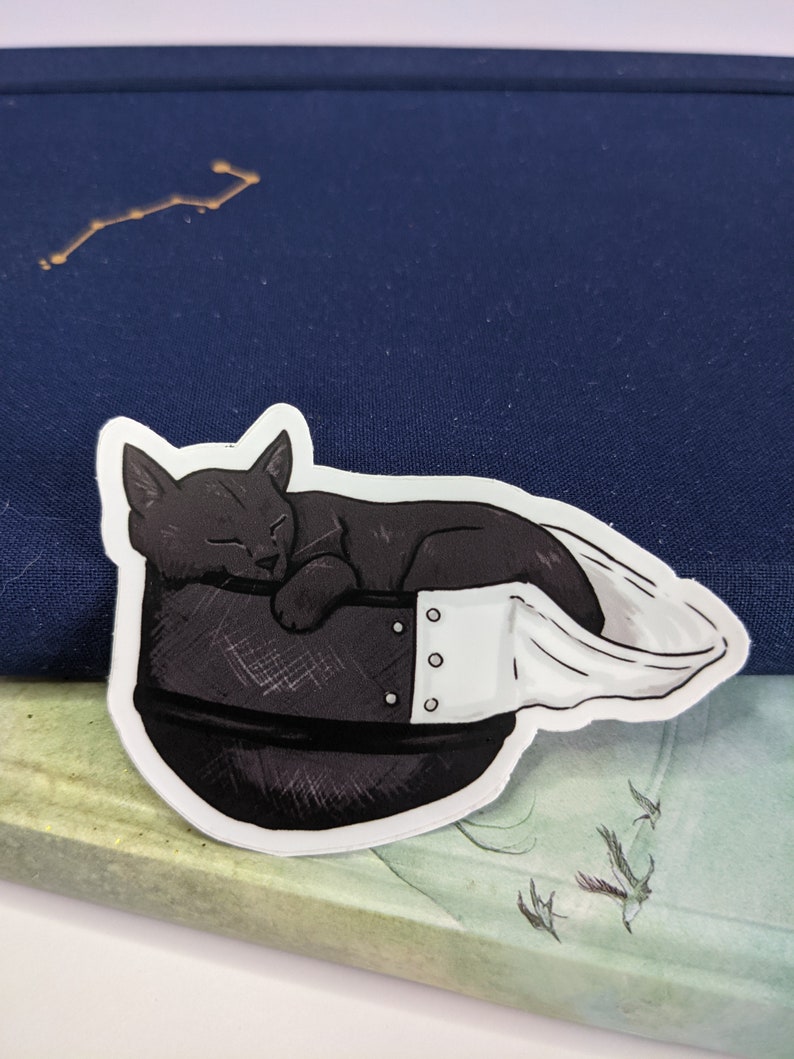 May include: A black cat sticker with white accents, sleeping inside a black and white helmet. The sticker is on a blue and green background.