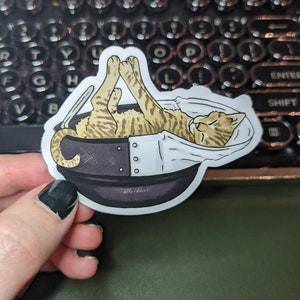 May include: A sticker of a tabby cat sleeping in a teacup. The cat is curled up in the cup with its paws in the air. The teacup is white with a black rim and a black handle.