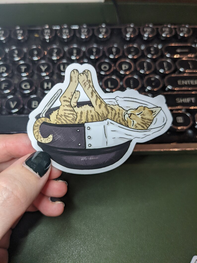 May include: A sticker of a tabby cat sleeping in a teacup. The cat is curled up in the cup with its paws in the air. The teacup is white with a black rim and a black handle.