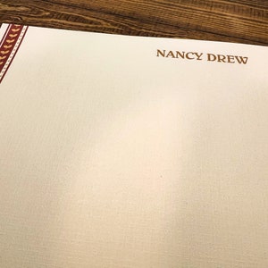 May include: A white stationery sheet with a red and gold border. The text "NANCY DREW" is printed in brown at the top of the sheet.