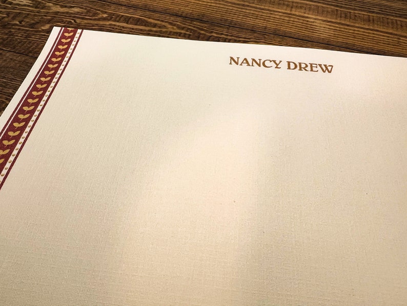 May include: A white stationery sheet with a red and gold border. The text "NANCY DREW" is printed in brown at the top of the sheet.