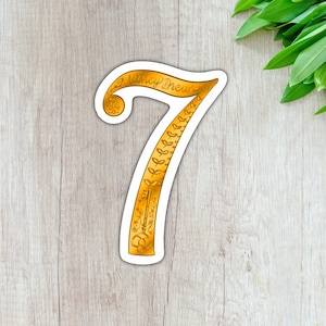 May include: Gold sticker with the number 7 in a decorative font. The number is outlined in white and has a floral design. The text "Nancy Drew" is written on the top of the number.