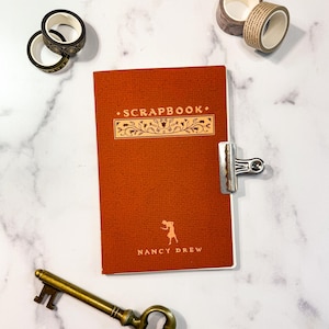 May include: A red scrapbook with the title "SCRAPBOOK" and the text "NANCY DREW" in gold lettering. The scrapbook has a floral design and a silhouette of a woman.