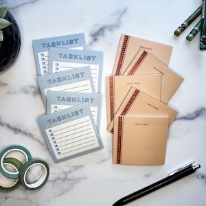 May include: An assortment of stationery items, including light blue task list notepads with the word "TASKLIST" printed on them, and tan "Nancy Drew" notebooks with a red decorative border. Also included are decorative tape rolls and a black pen.