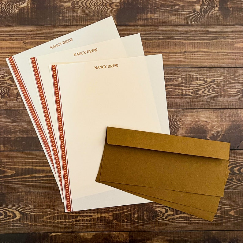 May include: Three sheets of white stationery with a red and gold border and the text "NANCY DREW" printed on each sheet.  Three brown envelopes are stacked on top of the stationery.