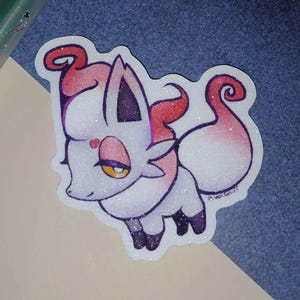 Puede incluir: Una pegatina con un zorro de dibujos animados con pelaje blanco, detalles rosas y un corazón. El zorro tiene un acabado brillante y está sobre un fondo azul. Se ven otros artículos de papelería en el fondo.