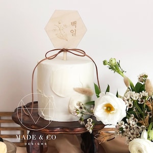 May include: A white cake with a hexagonal wooden cake topper that says "백일" in Korean. The cake is decorated with white frosting and a brown ribbon. The cake is on a wooden cake stand.
